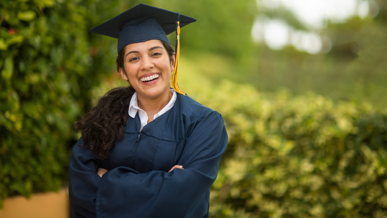 A woman in a graduation gown and cap smiles proudly at the camera