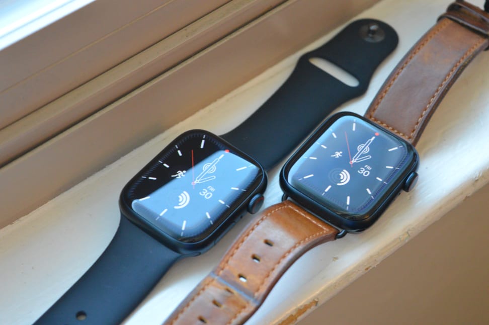 Two Apple Watches sit on a white ledge with their displays showing, one with a black band and the other with a brown leather band.