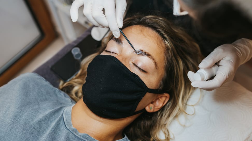 A person wearing a black face mask lays back with their eyes closed as a beautician runs a spoolie through their eyebrows.