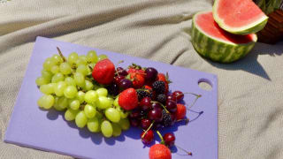 一个淡紫色的塑料切菜板，上面放着新鲜的水果，放在野餐毯上。