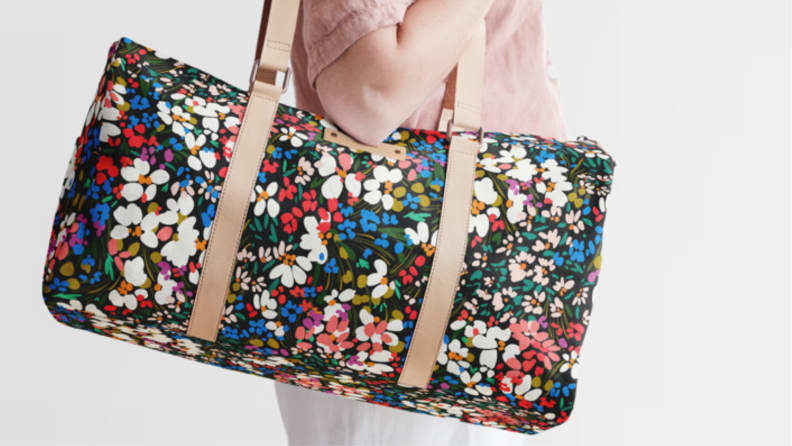 An image of a person holding the straps of a colorfully printed floral weekender bag.