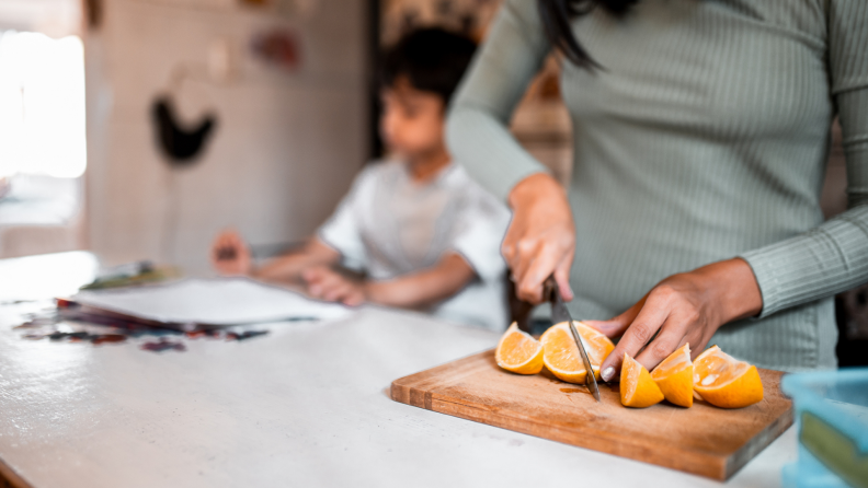 Person cutting up oranges on cutting board in kitchen next to child.