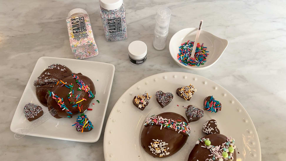 Chocolate eggs and hearts decorated with sprinkles sit on white plates, surrounded by jars and bowls of colorful sprinkles.