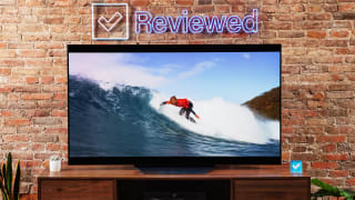 The LG B2 sits on a wooden table in front of a brick wall while it displays the image of a surfer.