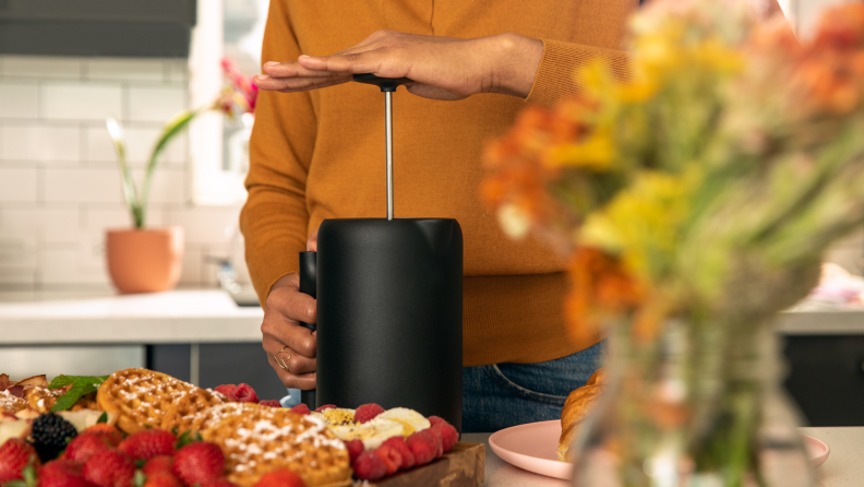 A person is plunging down a French press to make coffee in a kitchen. A plate of Belgian waffles and strawberries are on display as well.