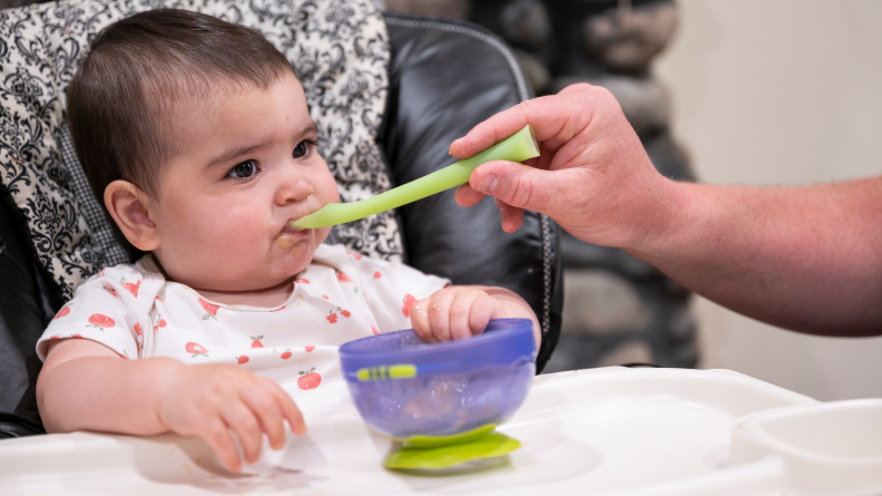 A baby sitting in a high chair being fed with a spoon by a parent.