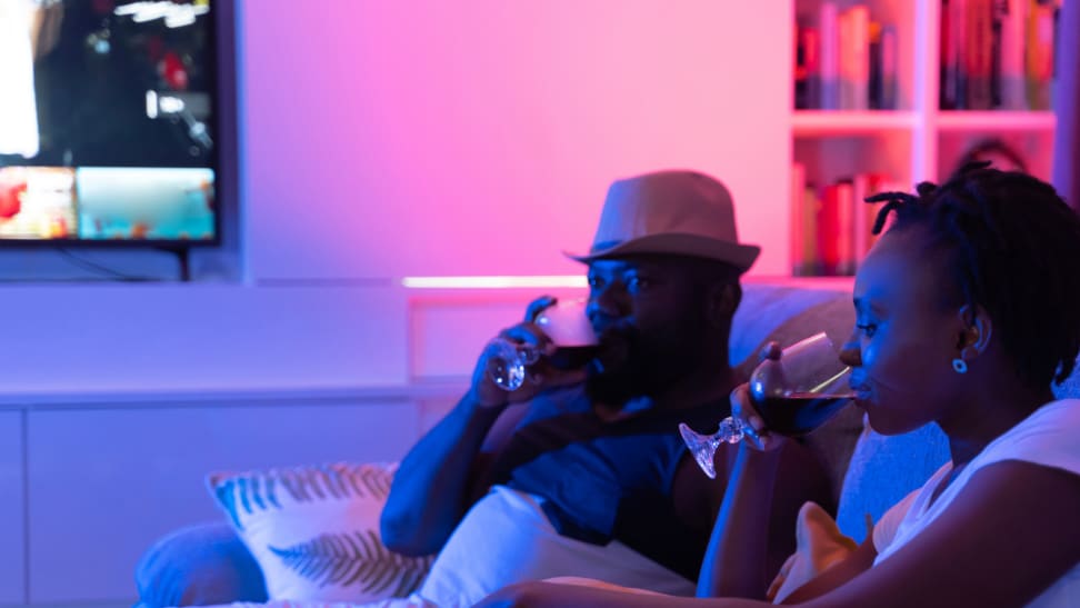 Two people sitting on a couch drinking out of glasses with colorful, ambient pink and blue lighting in the background