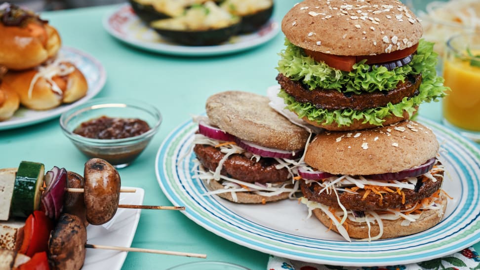 A spread with vegan burgers, skewers, and other out-of-focus plates with food on a blue tablecloth