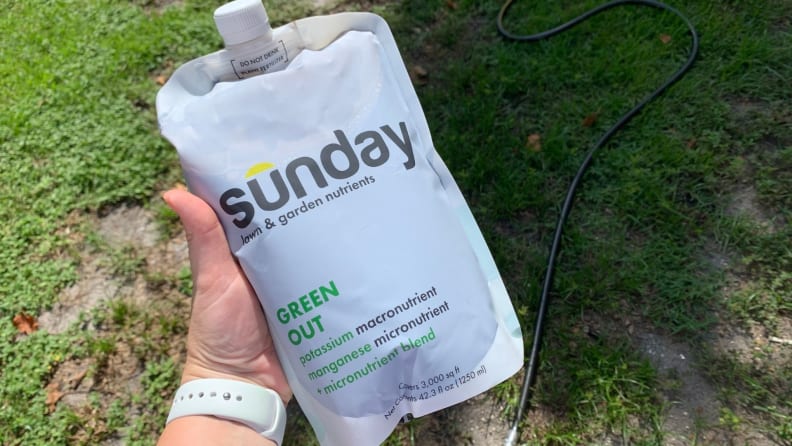 A woman wearing a white watch holds a Sunday lawn care nutrient pouch above the grass.