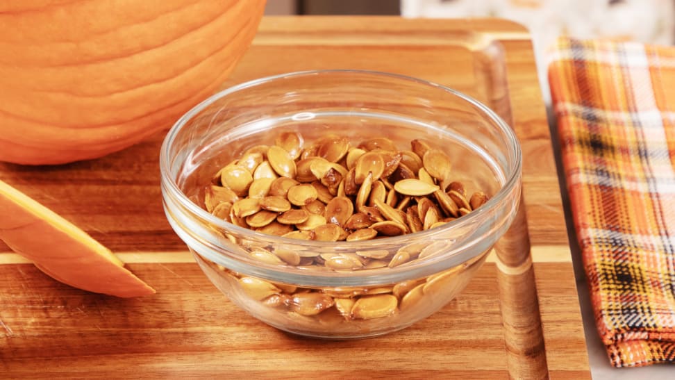 A clear bowl filled with pumpkin seeds on a wooden cutting board.