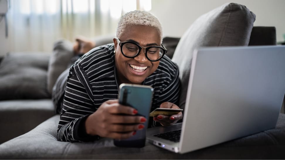 A person of color lays on a couch with a laptop and smartphone