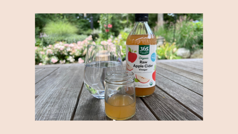 A glass of water, a small glass of cider vinegar, and a bottle of apple cider vinegar sit on an outdoor table.