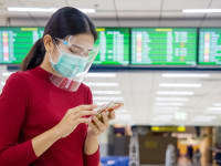 Women wearing red long-sleeved shirt, mask and face shield staring at the smart-phone she's holding while at the airport.