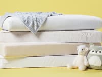 A stack of crib mattresses on a yellow background with two stuffed animals sitting on the floor next to them.