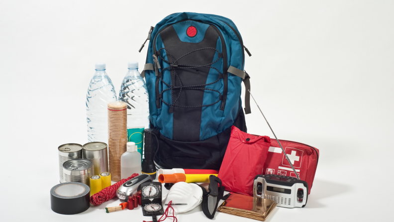 A survival kit backpack, sitting next to water bottles, a first aid kit, a radio, a flashlight, duct tape, canned goods, flares, a mask, and more smaller items.
