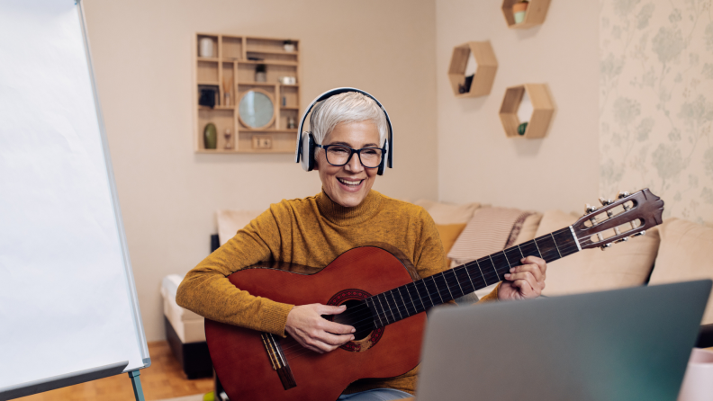 A person in an orange sweater smiles at their laptop, as if on a video call. They’re playing a nylon-stringed acoustic guitar.