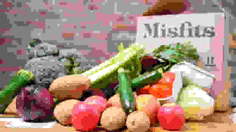 A pile of fresh produce such as potatoes, tomatoes, zucchini, celery, broccoli, red onion, lettuce, and apples sits in front of a Misfits Market box against a brick wall background.