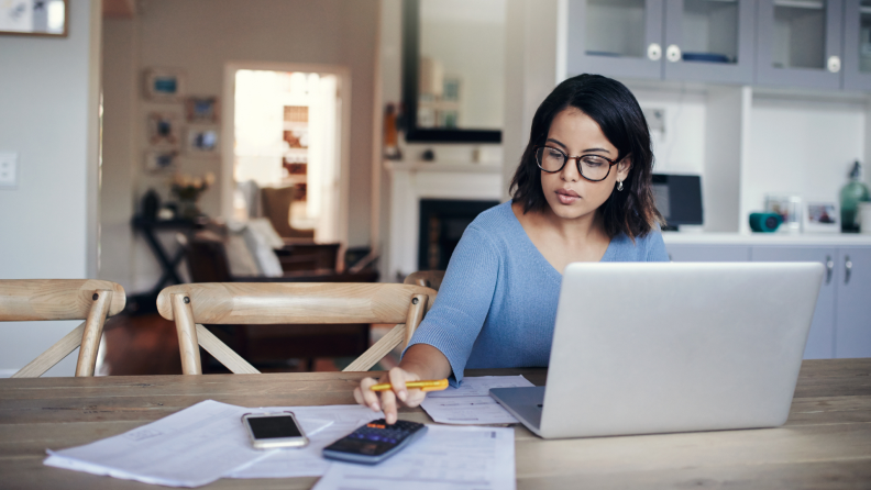 Person sitting at kitchen table in front of laptop computer using calculator to figure out finances.