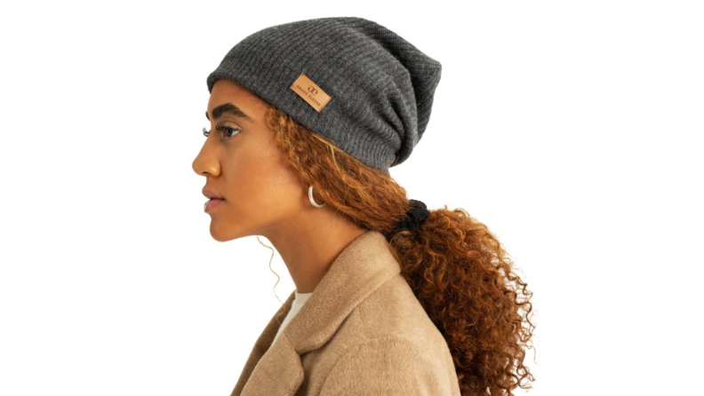 An image of a young Black woman with her hair pulled into a low ponytail, wearing a Grace Eyelae beanie in gray.