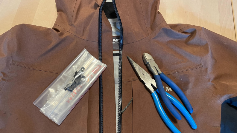 A Simms jacket in the color brown, with needle nose pliers and a zip repair kit on top of the jacket.