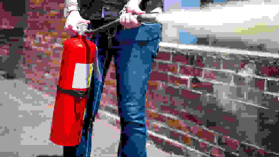 A waist-down shot of someone using a fire extinguisher in front of a brick wall.