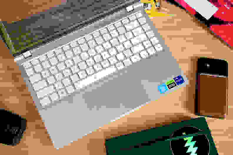 A top-down shot of a laptop with a white keyboard and silver chassis on top of a desk flanked by a few objects.