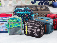 Eight colorful lunch coolers from brands like L.L. Bean, PackIt, Pottery Barn Kids, and Coleman sit on a kitchen counter.