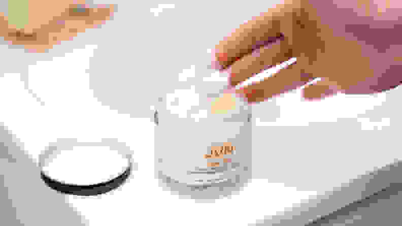 A jar of hair serum with a hand picking up a dollop of the white cream.