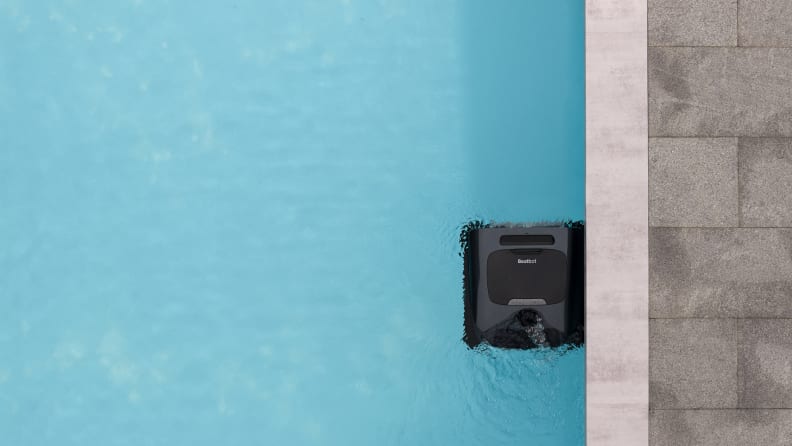 A black, square robot sits on the surface of the water by the edge of a swimming pool