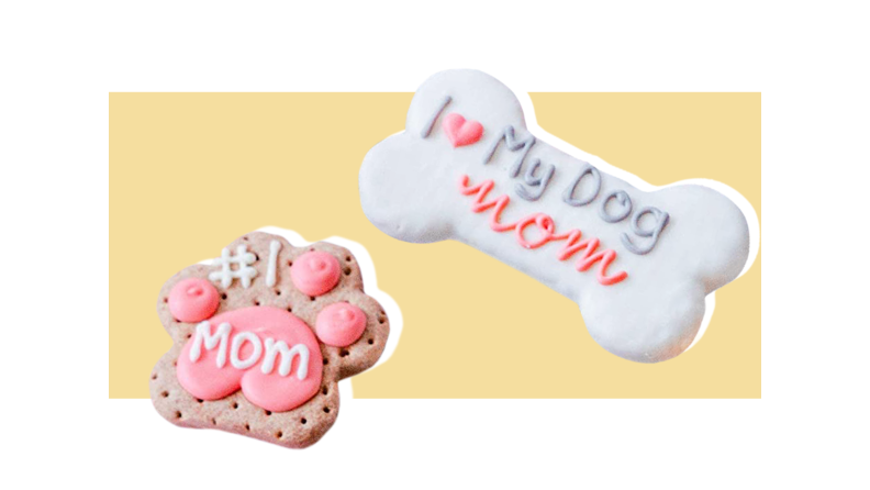 A paw shaped dog treat with pink icing and the words "#1 Mom" in white icing in the lower left corner, and a blue bone shaped treat with "I (heart) My Dog" in blue icing with a pink heart, and "Mom" below it in pink icing against a light gold background.