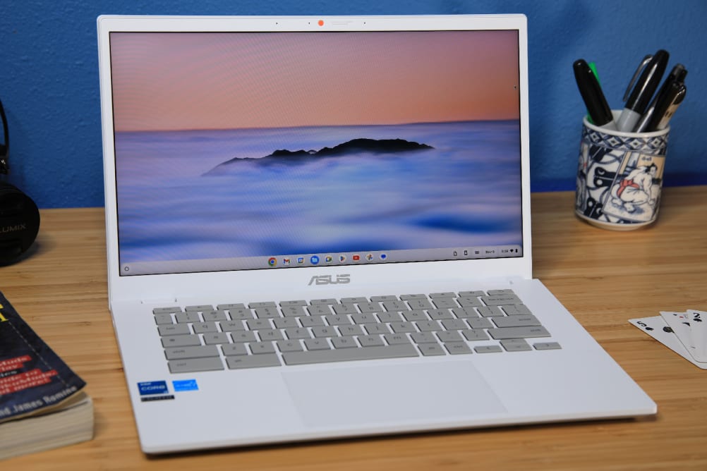 The Asus Chromebook Plus CX34 sitting open on a desk displaying a cloudy landscape with a cup of pens to the right and a paperback book to the left.