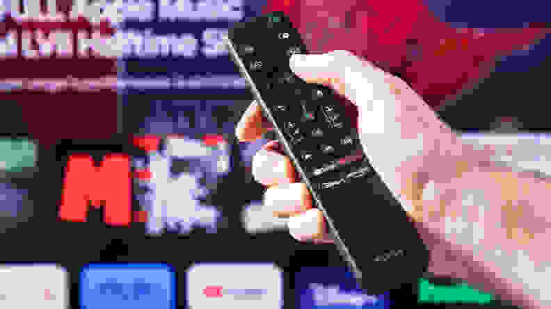 A person holding a Sony X90K LED TV remote with the Sony X90K LED TV's home screen, blurred in the background.
