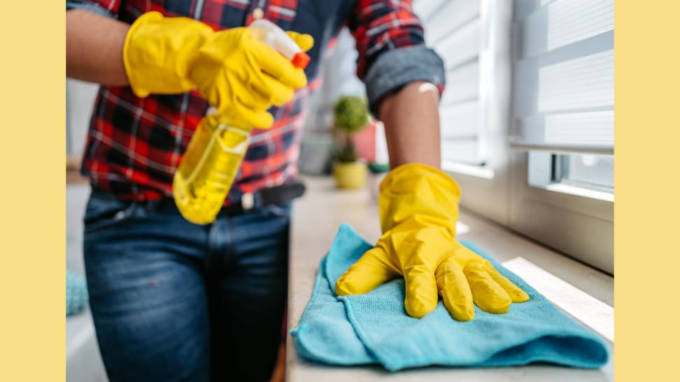 Person wearing yellow rubber gloves is dusting a window ledge with a blue rag and a yellow spray bottle.