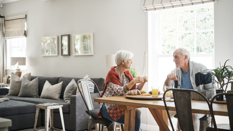 Senior couple sitting together having a discussion at dining room table in bright room surrounded by windows.