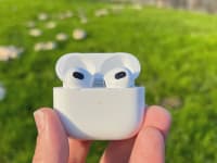 The all-white Apple AirPods (3rd) gen sit in their open case before a green pasture with a trail of mushrooms.
