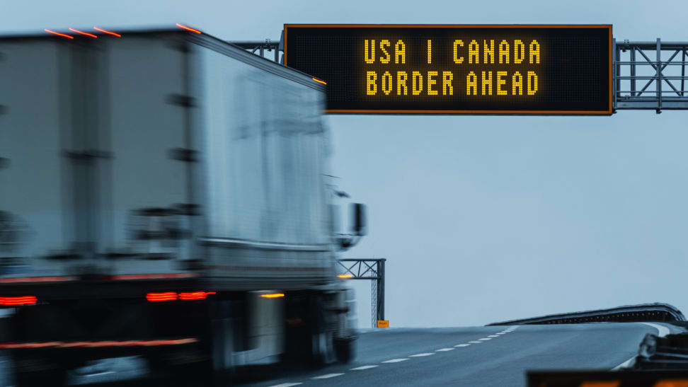 A truck drives a highway under a sign that reads USA Canada border