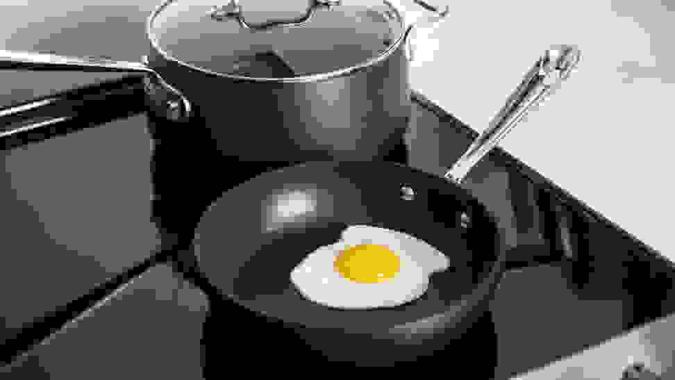 A perfect fried egg inside a nonstick pan, with a nonstick pot behind it, both sitting on a stovetop.