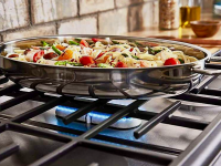 Close-up of a flavorful pasta dish in a silver pain. It sits on a gas stovetop with a blue flame glowing beneath it.