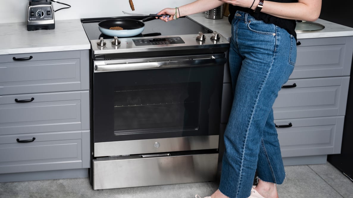 A person wearing jeans stands in front of an electric range cooking food in a pan on the stovetop.