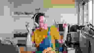 Woman wearing yellow shirt and overalls, sitting in living dancing to music while wearing headphones and holding smartphone.