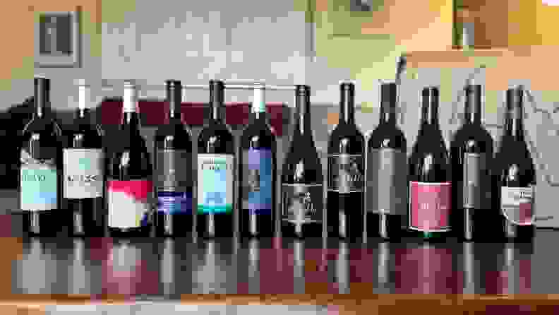 A row of 12 wine bottles on top of wooden table in front of cardboard box.