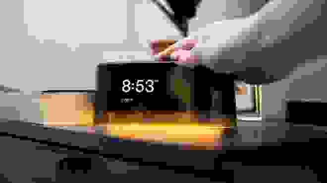 Person touching the top of glowing alarm clock next on bedside table.