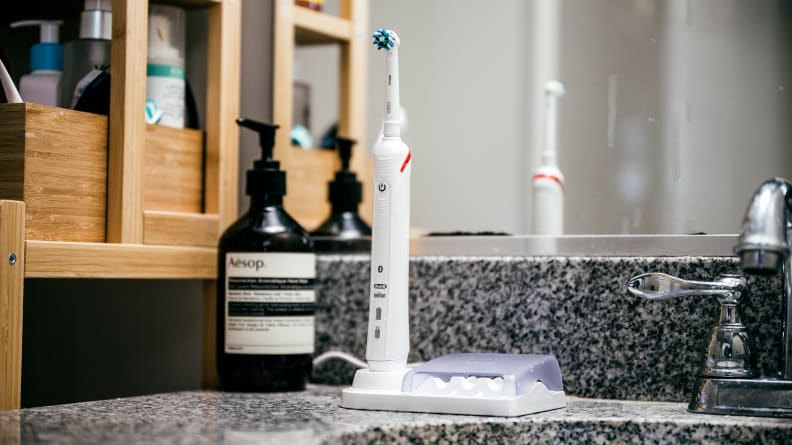 An Oral B Pro 3000 electric toothbrush sits on its charger on a dark countertop, next to a sink.