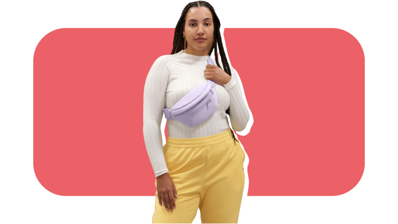 A model wearing a lavender Dagne Dover Ace fanny pack with a yellow pair of sweatpants and a long-sleeve top.