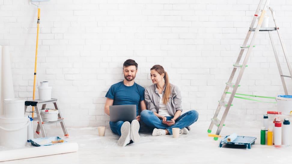 Two people sitting against a white wall surrounded by home improvements supplies.
