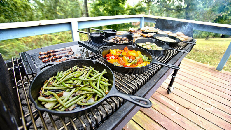 Several cast iron pans filled with cooking vegetables on a big outdoor grill.
