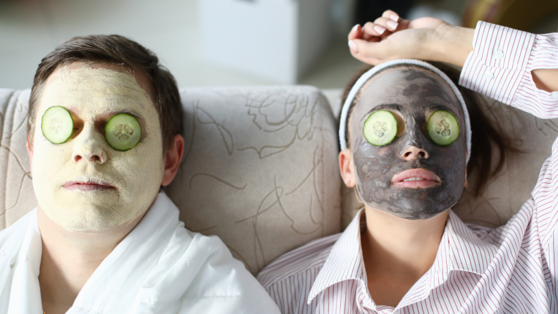 A couple luxuriates on the couch, each wearing a face mask with cucumbers over their eyes.