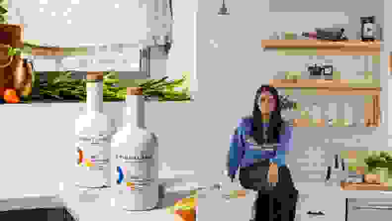 On left, two bottles of Brightland olive oil on white countertop. On right, Brightland Founder Aishwarya Iyer sitting on a white counter surrounded by Brightland products