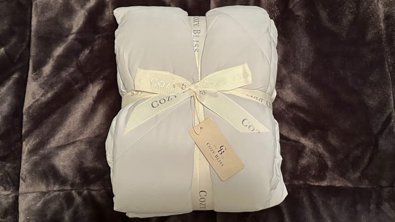 Gray Cozy Bliss comforter folded into a square cushion shape tied together with a white ribbon, laying on a brown fuzzy blanket.
