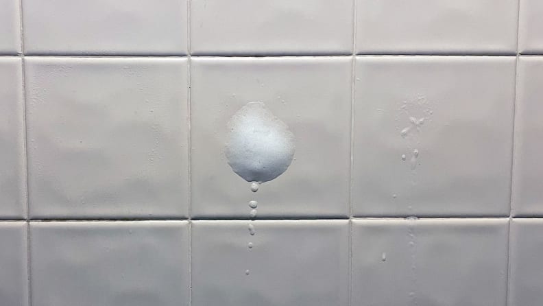 A close-up of three tiles, all of which have been sprayed with shower cleaner. The leftmost shows a barely-visible mist pattern that coats the tilie evenly. The second shows some mild foaming action puffing up in the tile's center. The rightmost shows residue from the spray's initial point of impact, but almost all of the spray has dripped down at this point.